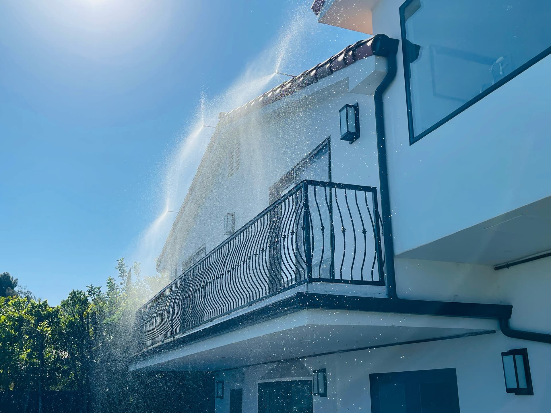Eave sprayers running along the underside of a two-story home’s overhang, throwing a wide curtain of water down across the wall and balcony.
