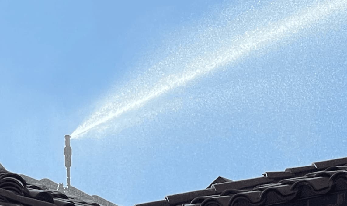 A FireShield roof rotor mounted on a tile ridge, throwing a long jet of water across the roof against a clear blue sky.