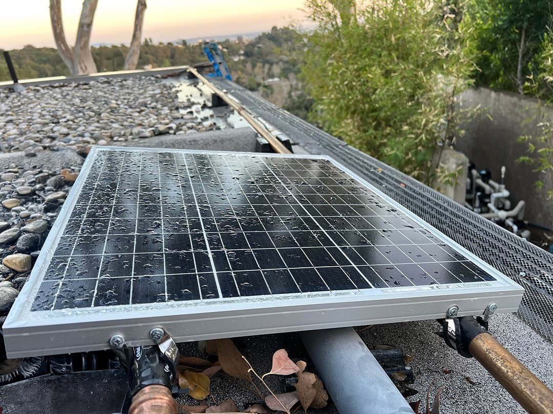 A solar panel mounted on a flat gravel roof beside the system's copper supply line at sunset, keeping the backup battery charged.